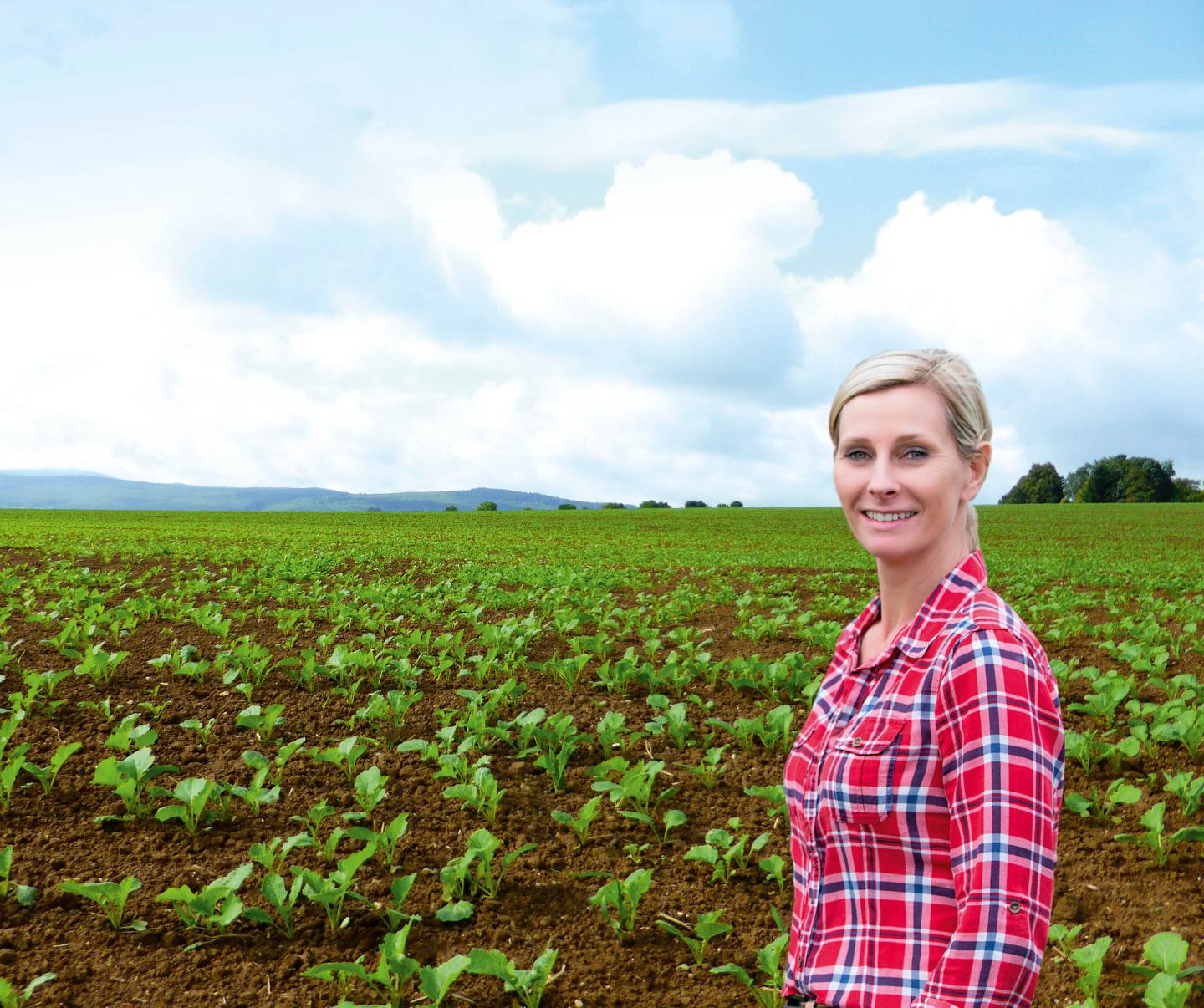 Lächelnde Frau in rot-blauem Karohemd auf grünem Rapsfeld mit Bergen und Himmel im Hintergrund.