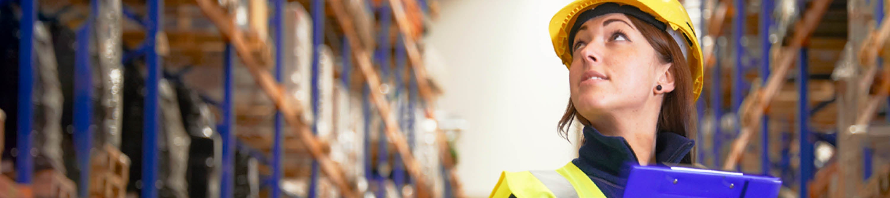 Frau mit gelbem Helm und blauem Klemmbrett in Lagerhalle